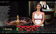 Ein weiblicher Croupier sitzt neben einem Roulettetisch. Im Hintergrund sitzt ein Blackjack-Dealer.