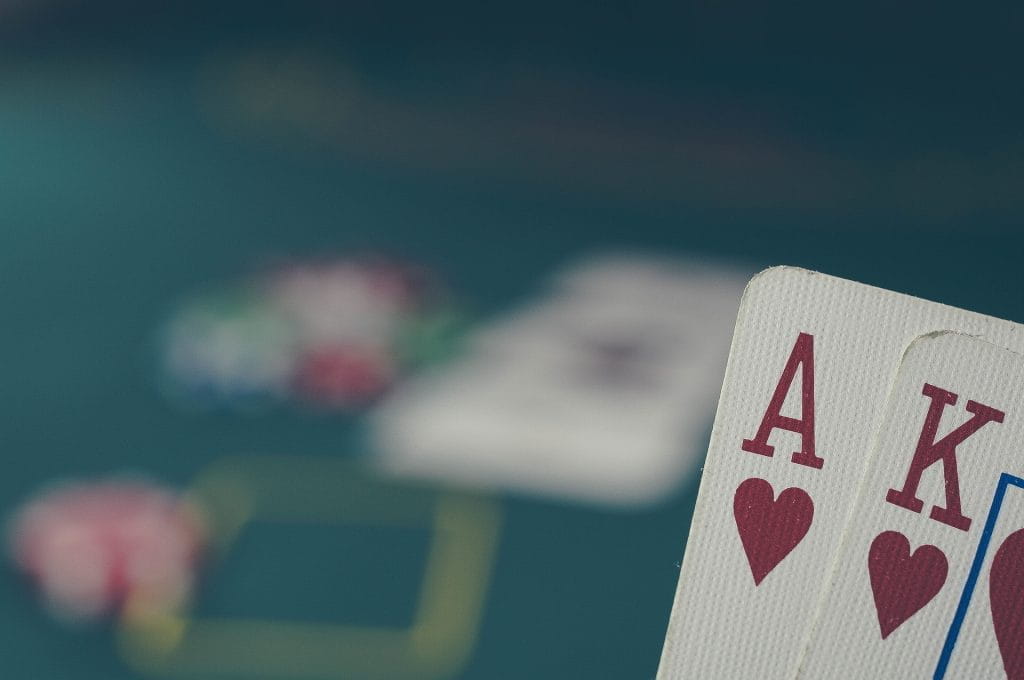 Die Pokerkarten Ass Herz und König Herz werden vor einem Pokertisch gehalten.