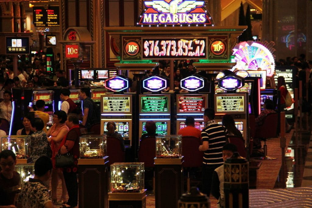 Zahlreiche leuchtende Spielautomaten in einem Casino in Macau; dazwischen Menschen beim Spielen.