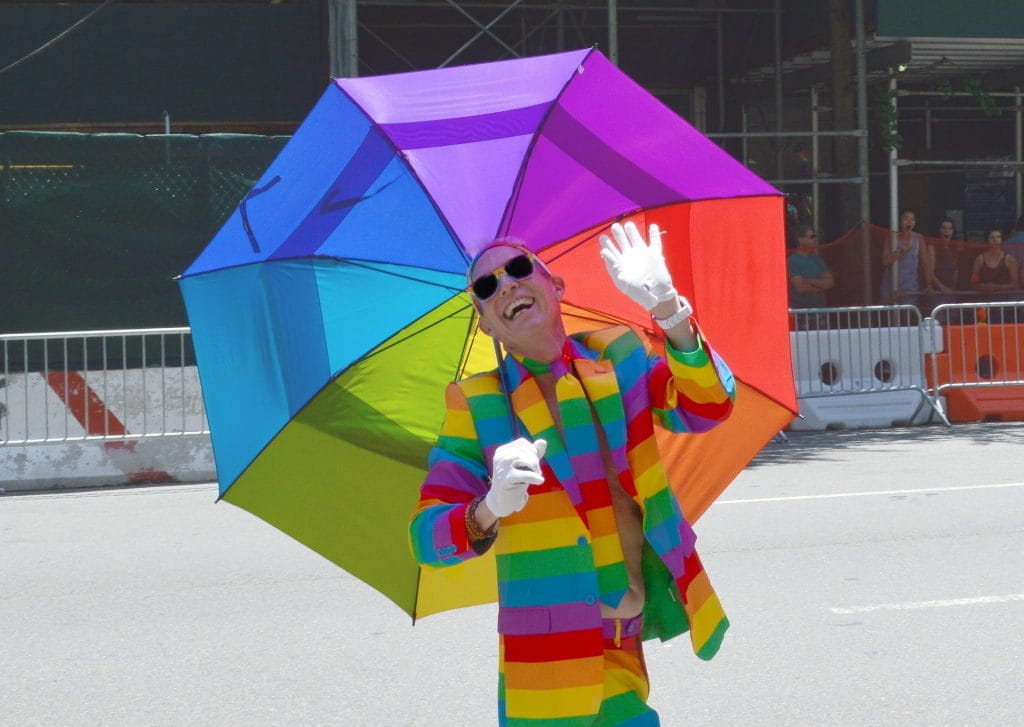 Mann in Regenbogen-Anzug hält auf Straße einen Regenbogenschirm lachend und winkend in die Kamera.