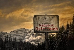 Heruntergekommenes Schild mit Aufschrift Republik Österreich vor dunklem Himmel und bewaldeten Bergen.