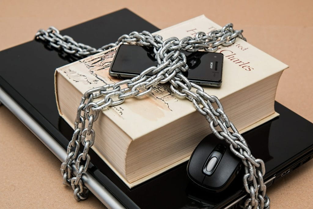 Ein Laptop, ein Gesetzesbuch, eine PC-Maus und ein Smartphone sind von einer Eisenkette umschlossen.