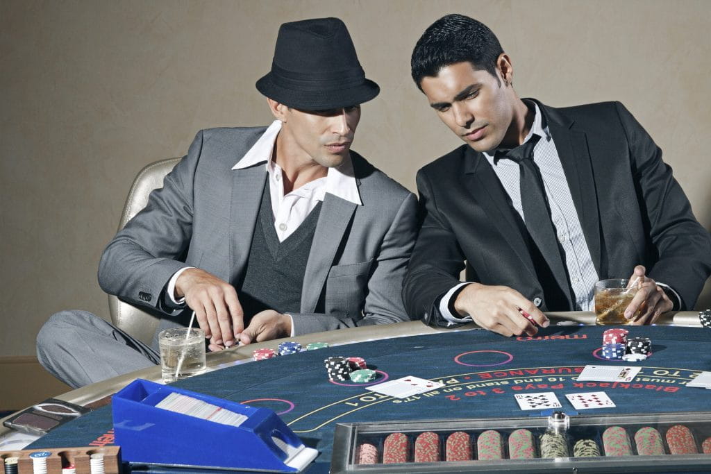 Zwei Männer im Anzug sitzen an einem Blackjack-Tisch und diskutieren über ihre Karten.
