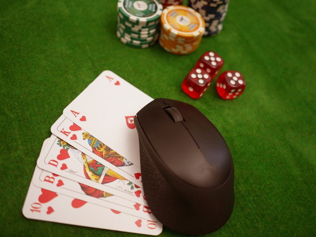 Pokerchips, Würfel sowie fünf Spielkarten liegen neben einer Maus für den Computer auf einem Tisch.
