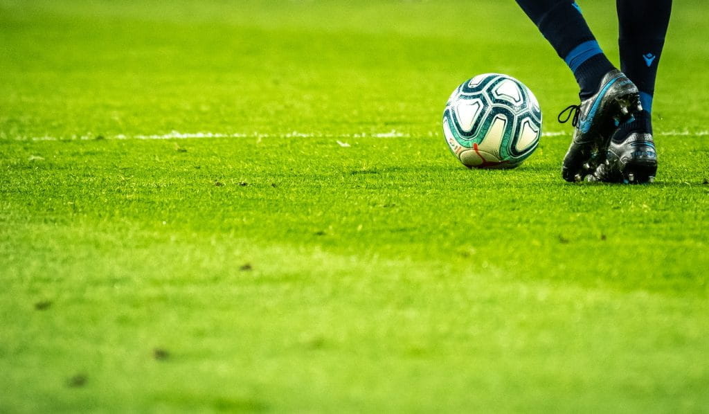 Ein Fußballspieler mit einem Ball am Fuß steht auf einem Rasenplatz.