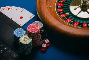 Jetons auf einem Laptop samt Roulette-Kessel und Spielkarten.