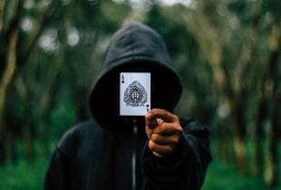 Ein Mann mit schwarzer Kapuze hält eine Spielkarte vor sein Gesicht.