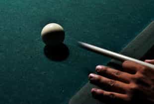 Eine Person schlägt auf einem Snooker-Tisch eine weiße Kugel mit einem Queue an.