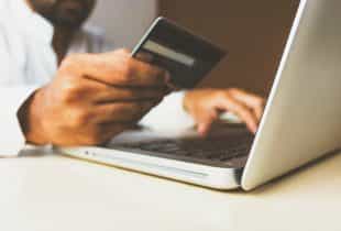 Eine Person sitzt an einem Laptop mit einer Kreditkarte in der rechten Hand.