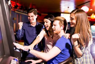 Zwei Personen spielen an einem Casinoautomaten, während im Hintergrund Lichter und Geräusche einer belebten Spielhalle wahrnehmbar sind.