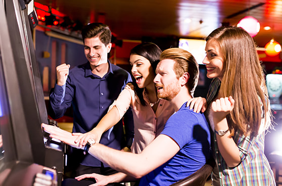 Zwei Personen spielen an einem Casinoautomaten, während im Hintergrund Lichter und Geräusche einer belebten Spielhalle wahrnehmbar sind.