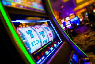 Mehrere unscharfe Glücksspielautomaten stehen in einem dunklen Raum. Im Vordergrund ist ein Automat mit leuchtendem Bildschirm zu erkennen.
