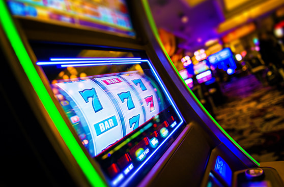 Mehrere unscharfe Glücksspielautomaten stehen in einem dunklen Raum. Im Vordergrund ist ein Automat mit leuchtendem Bildschirm zu erkennen.