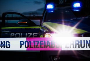 Ein Polizeiauto mit eingeschaltetem Blaulicht steht nachts vor einem abgesperrten Bereich, der mit Flatterband markiert ist.