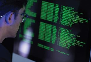 Ein Computermonitor zeigt komplexe Zeilen von Programmcode in grüner Schrift auf dunklem Hintergrund, die von einer Person analysiert werden.