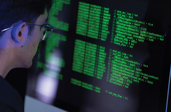 Ein Computermonitor zeigt komplexe Zeilen von Programmcode in grüner Schrift auf dunklem Hintergrund, die von einer Person analysiert werden.