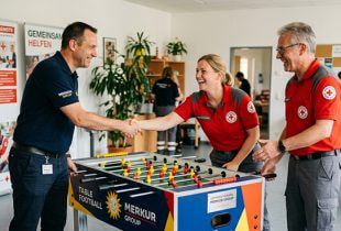 Zwei Vertreter des Deutschen Roten Kreuzes nehmen freudig einen neuen Kickertisch von einem Mitarbeiter der Merkur Group entgegen.